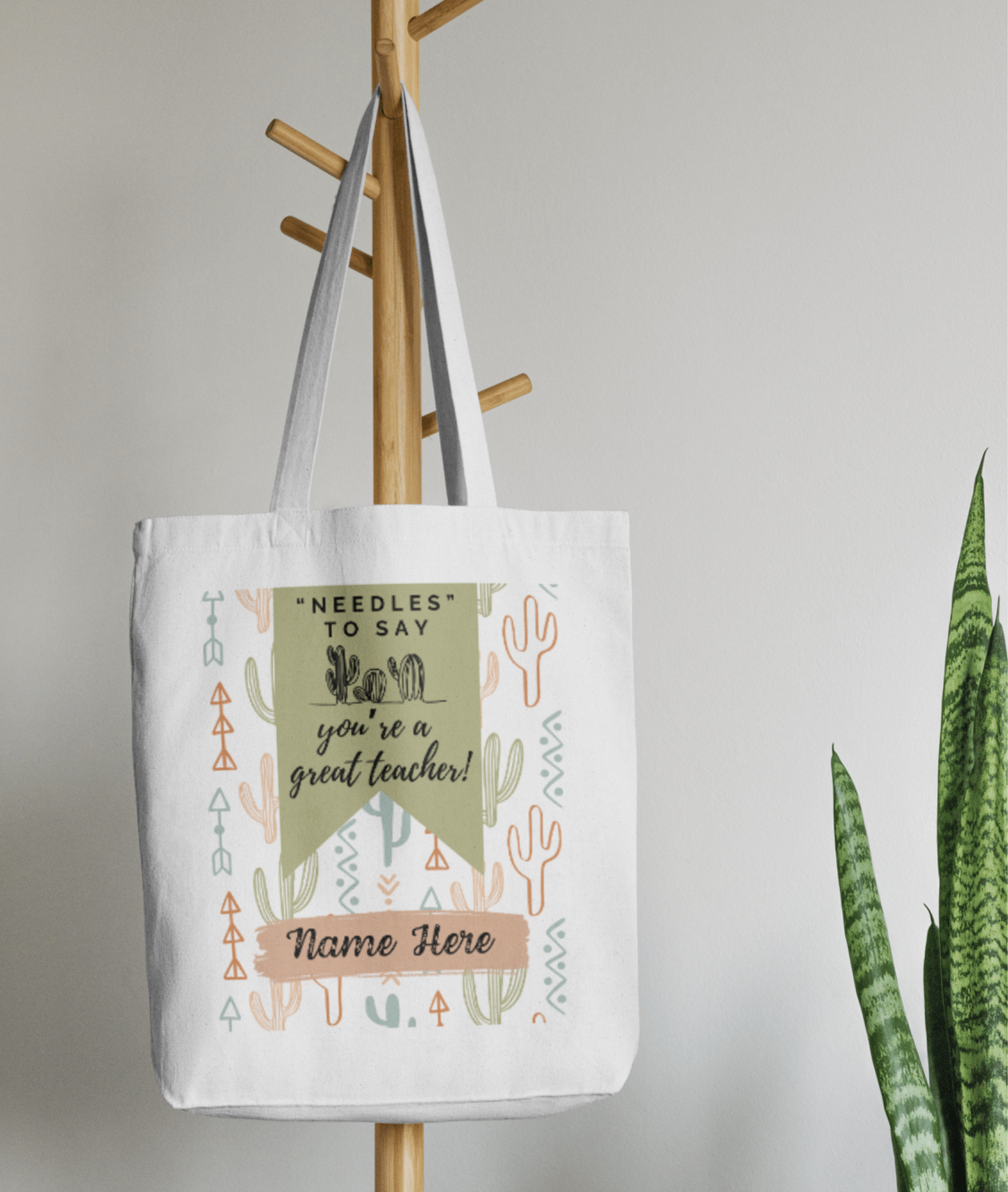 White cotton tote bag with pastel cactus and arrow pattern, green banner with the text "'Needles' to say you're a great teacher!" and customizable name section, hanging on a wooden coat rack.