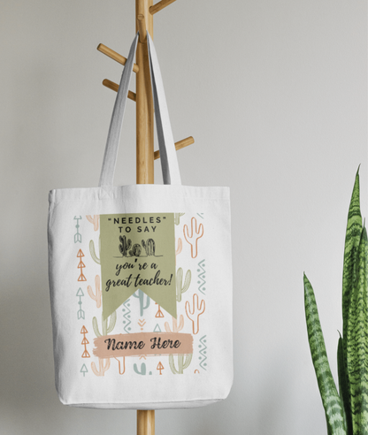 White cotton tote bag with pastel cactus and arrow pattern, green banner with the text "'Needles' to say you're a great teacher!" and customizable name section, hanging on a wooden coat rack.